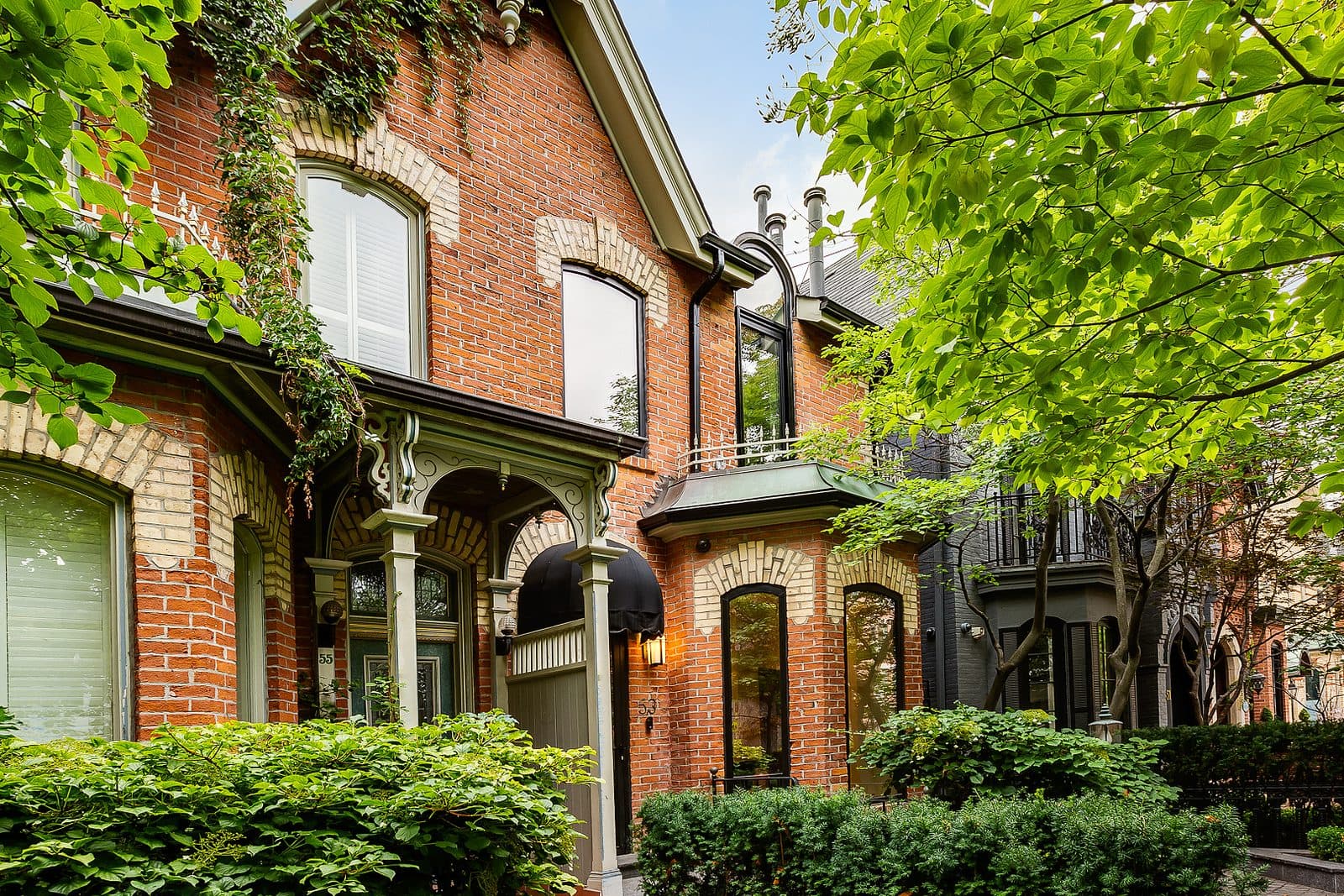 Two story red brick Victorian facade with cream brick banding, arched black framed windows, iron fence, and mature trees along the front walk.