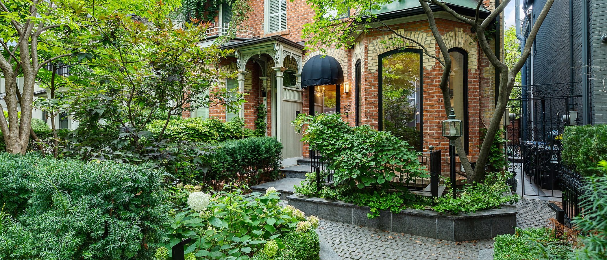 Stone paver walkway leading to a black glass front door beneath a black canopy awning, flanked by hydrangeas and clipped hedges.