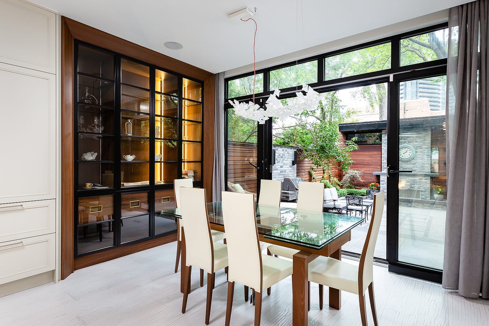 Dining room with glass table, upholstered chairs, and a walnut framed glass front cabinet lit from within, opening to a rear terrace.