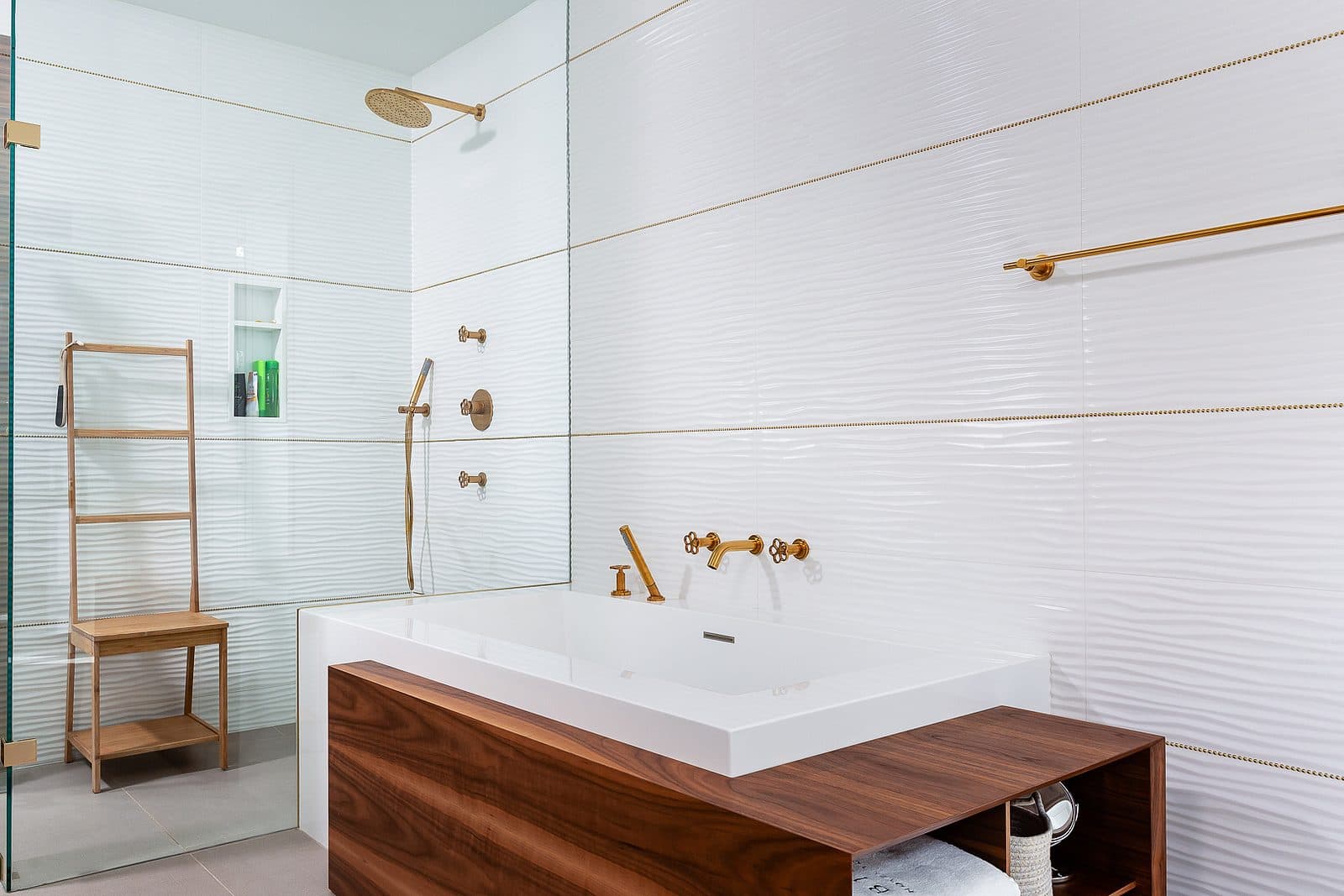 Freestanding tub in a walnut surround beneath a brass wall mounted filler, with a glass shower and brass rain head beyond.