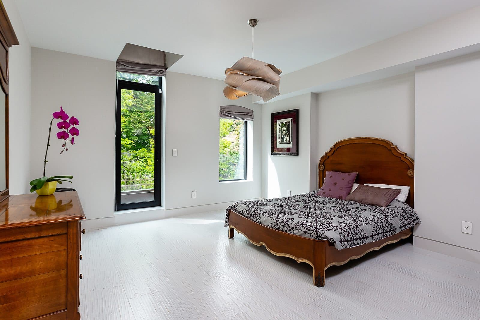 Bright bedroom with a carved wood antique bed frame, matching dresser, orchid, and a tall window overlooking trees.