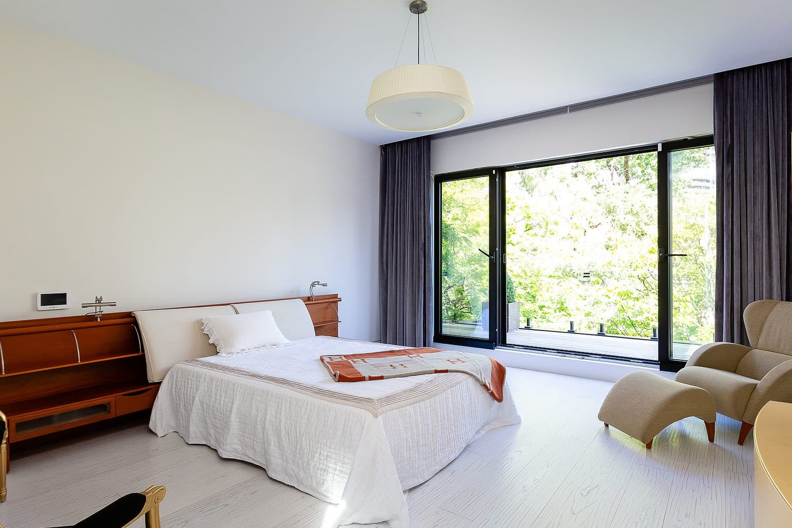 Bedroom with a wood headboard bed, drum pendant light, and full height glass doors opening onto a tree lined balcony.