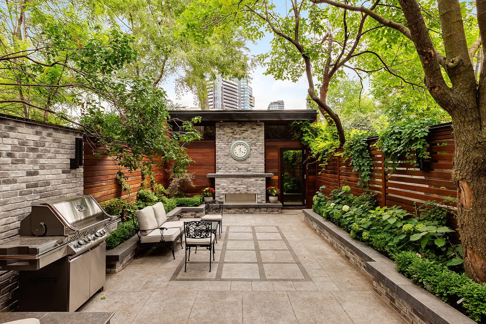 Stone paved rear patio with a built-in stone fireplace, wall clock, wrought iron seating, and stainless outdoor kitchen framed by wood fencing and trees.