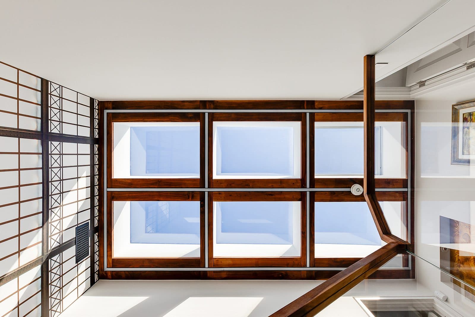 Upward view of a walnut framed skylight window grid over a stairwell, with steel lattice paneling and a stair rail in the foreground.