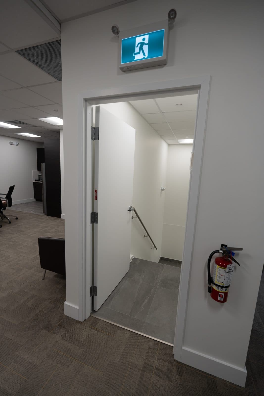 Open plan office edge with a leather lounge chair, stairwell door propped open, and a wall mounted fire extinguisher.