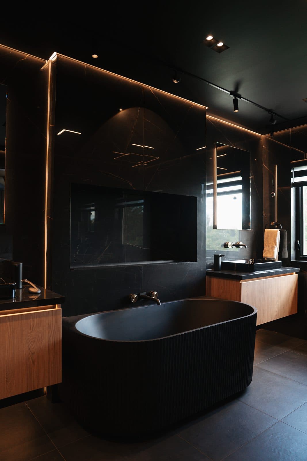 Fluted black freestanding tub beside a warm oak vanity in the dark ensuite