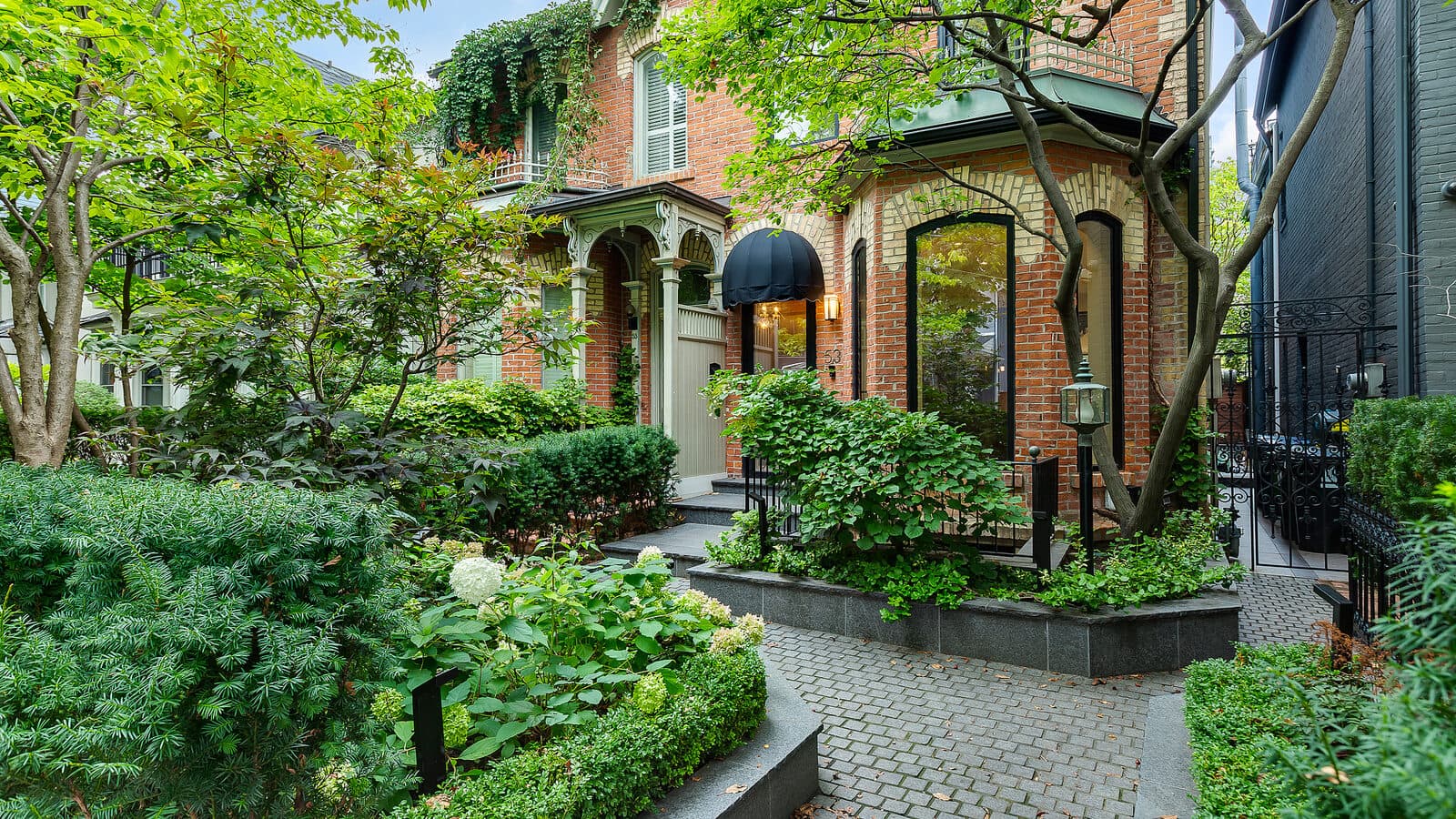 Stone paver walkway leading to a black glass front door beneath a black canopy awning, flanked by hydrangeas and clipped hedges