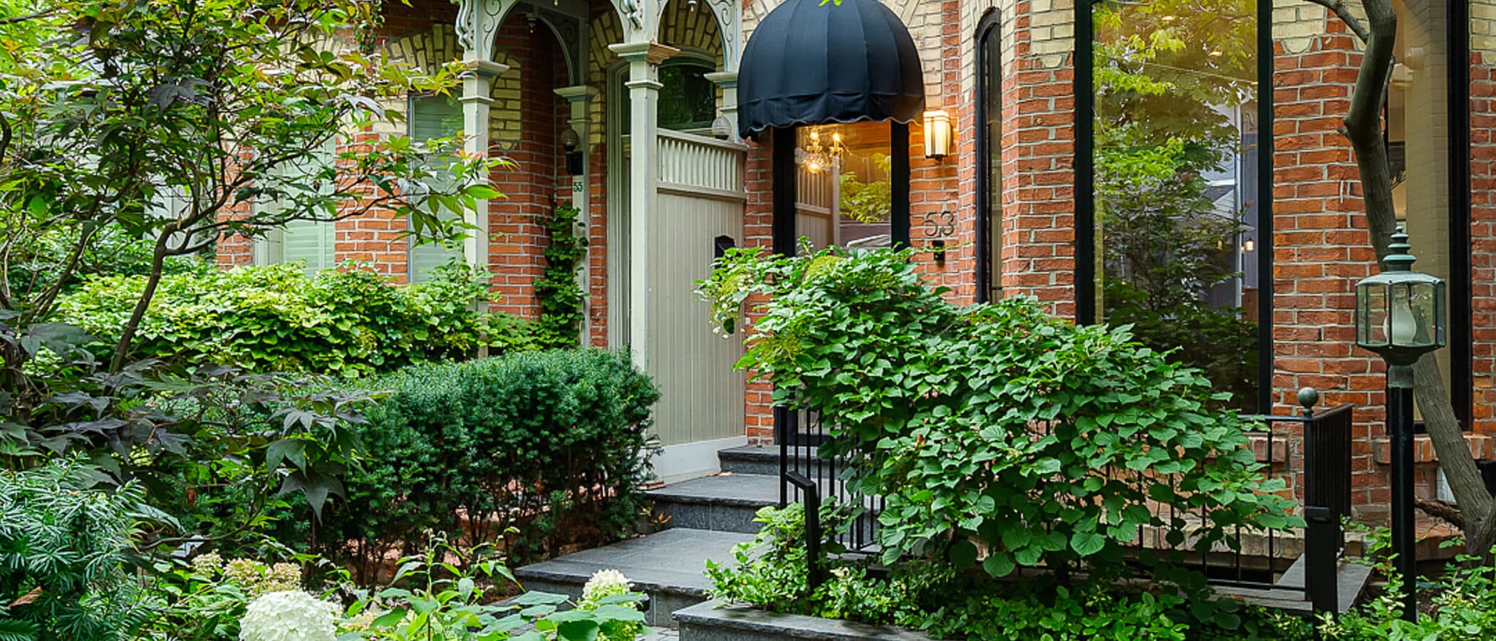 Stone paver walkway leading to a black glass front door beneath a black canopy awning, flanked by hydrangeas and clipped hedges