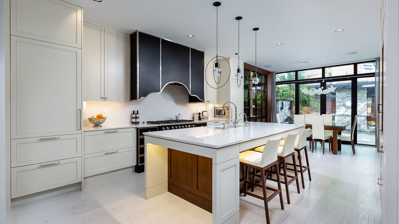 Open concept kitchen with white cabinetry, black paneled range hood, waterfall island, and dining table beyond glass doors to the garden