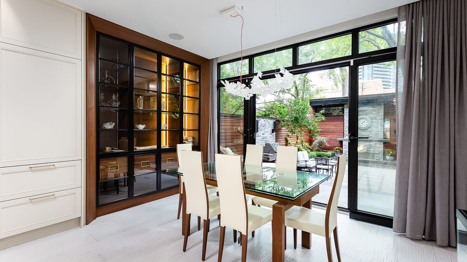 Dining room with glass table, upholstered chairs, and a walnut framed glass front cabinet lit from within, opening to a rear terrace