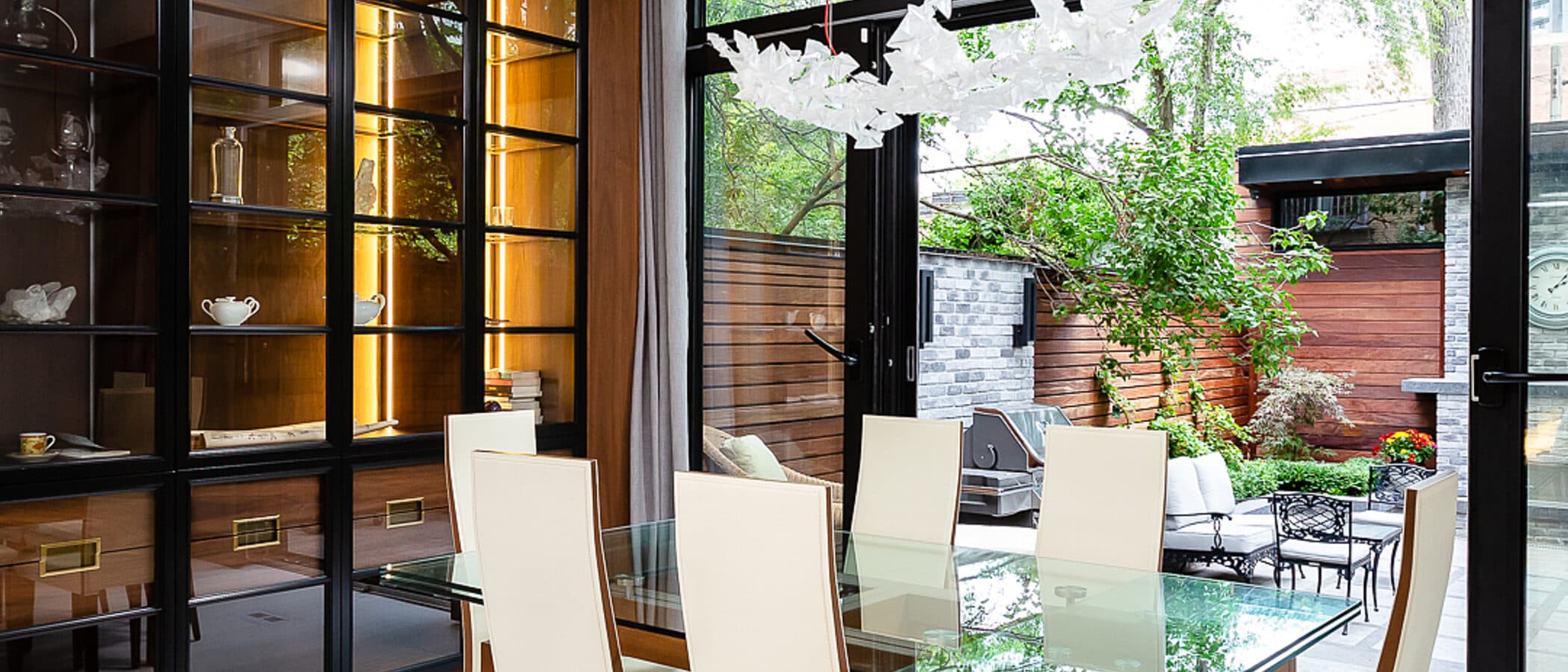 Dining room with glass table, upholstered chairs, and a walnut framed glass front cabinet lit from within, opening to a rear terrace