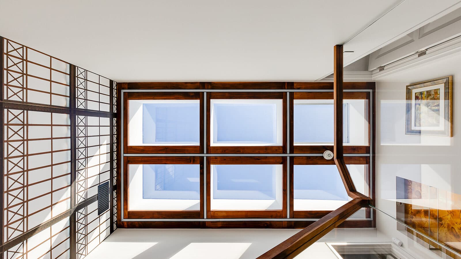 Upward view of a walnut framed skylight window grid over a stairwell, with steel lattice paneling and a stair rail in the foreground