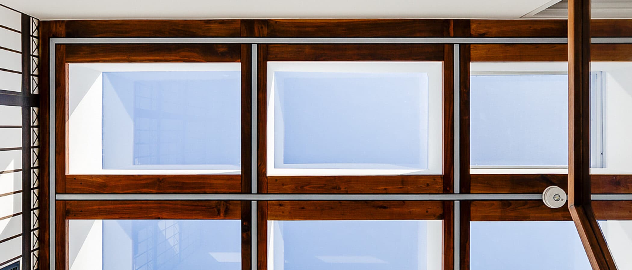Upward view of a walnut framed skylight window grid over a stairwell, with steel lattice paneling and a stair rail in the foreground