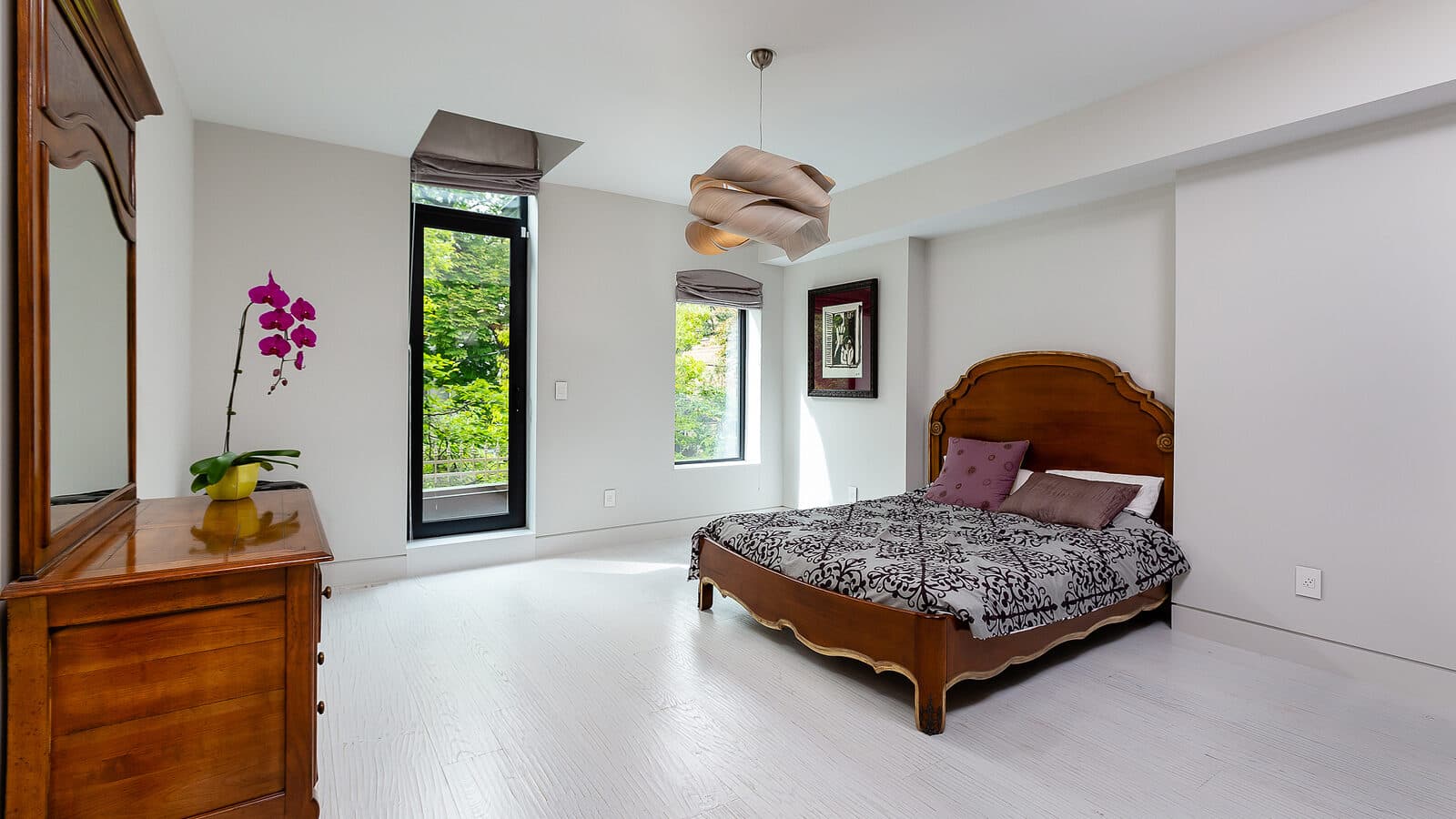 Bright bedroom with a carved wood antique bed frame, matching dresser, orchid, and a tall window overlooking trees
