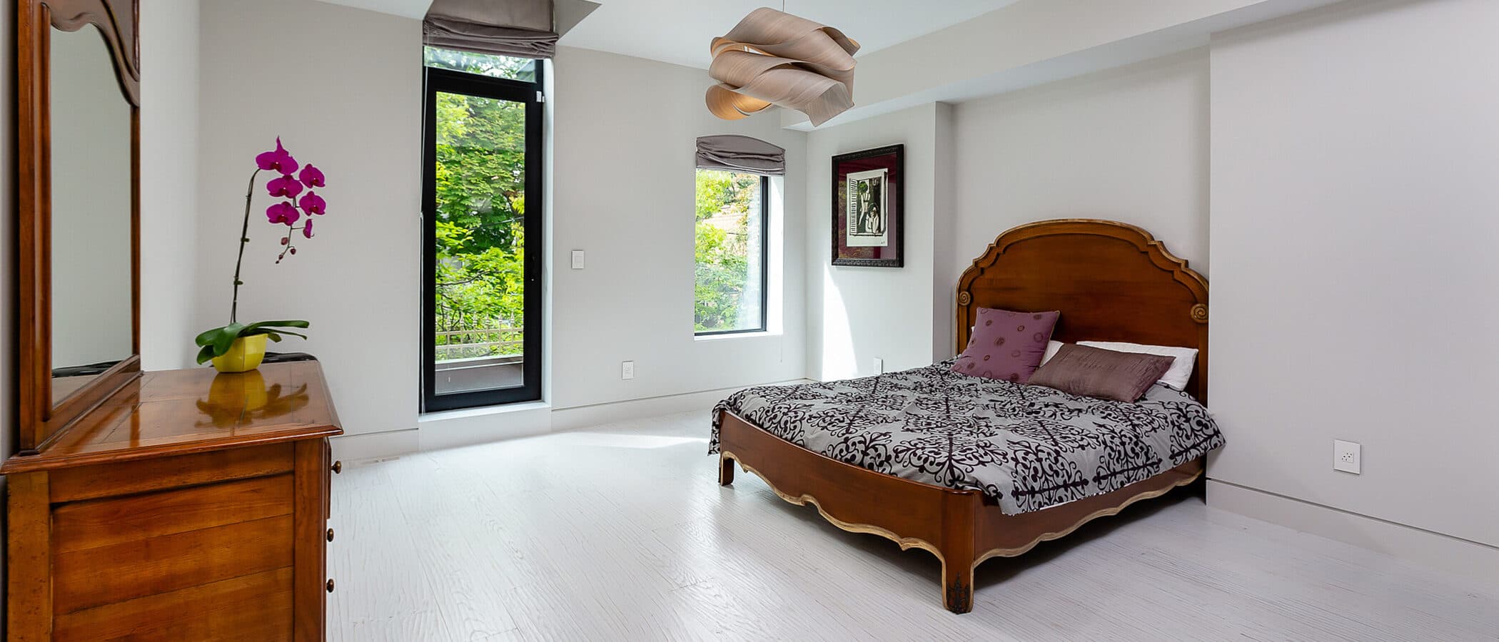 Bright bedroom with a carved wood antique bed frame, matching dresser, orchid, and a tall window overlooking trees