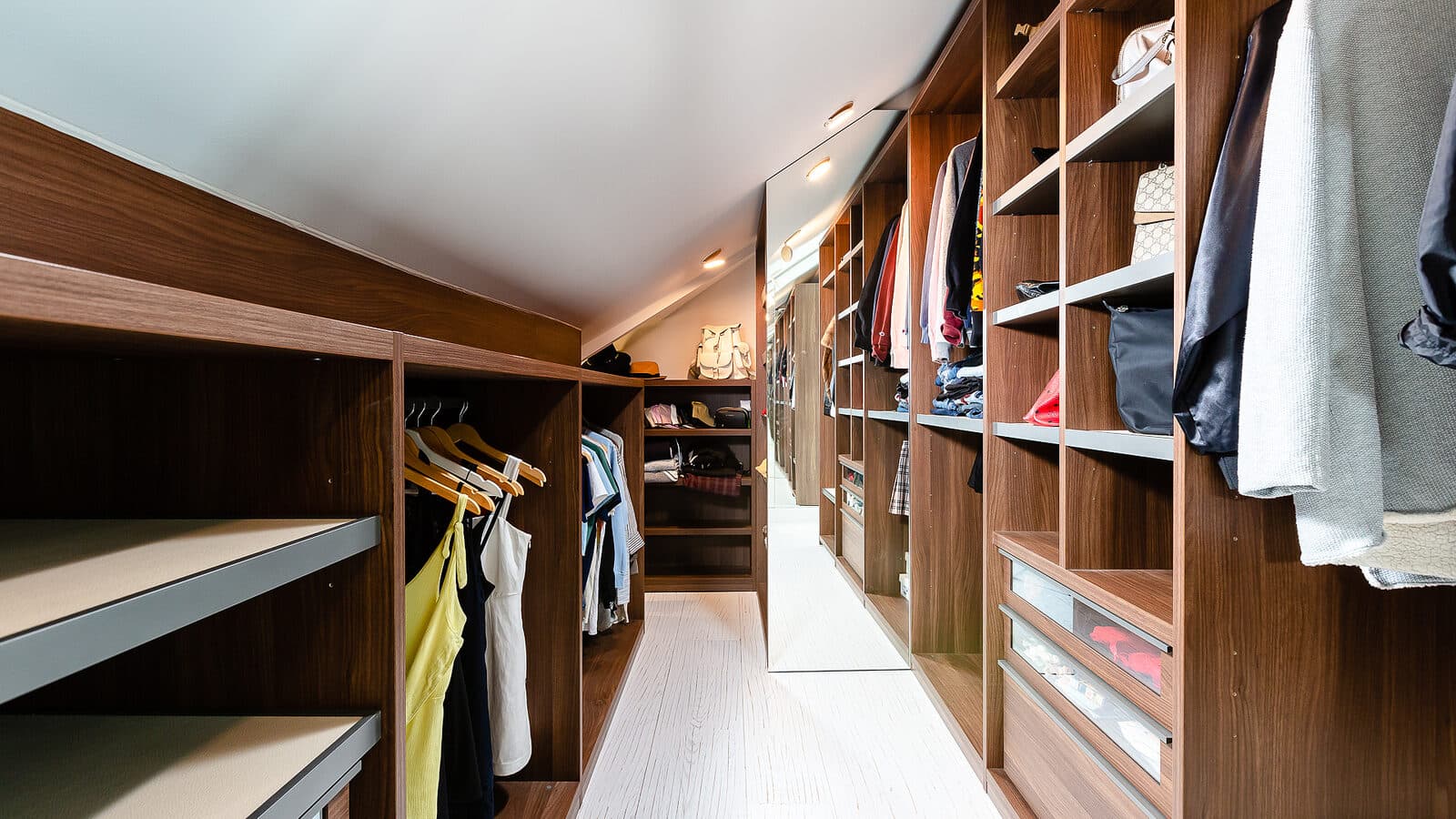 Walnut walk in closet with open hanging bays, built in drawers, and a mirrored end wall under a sloped ceiling