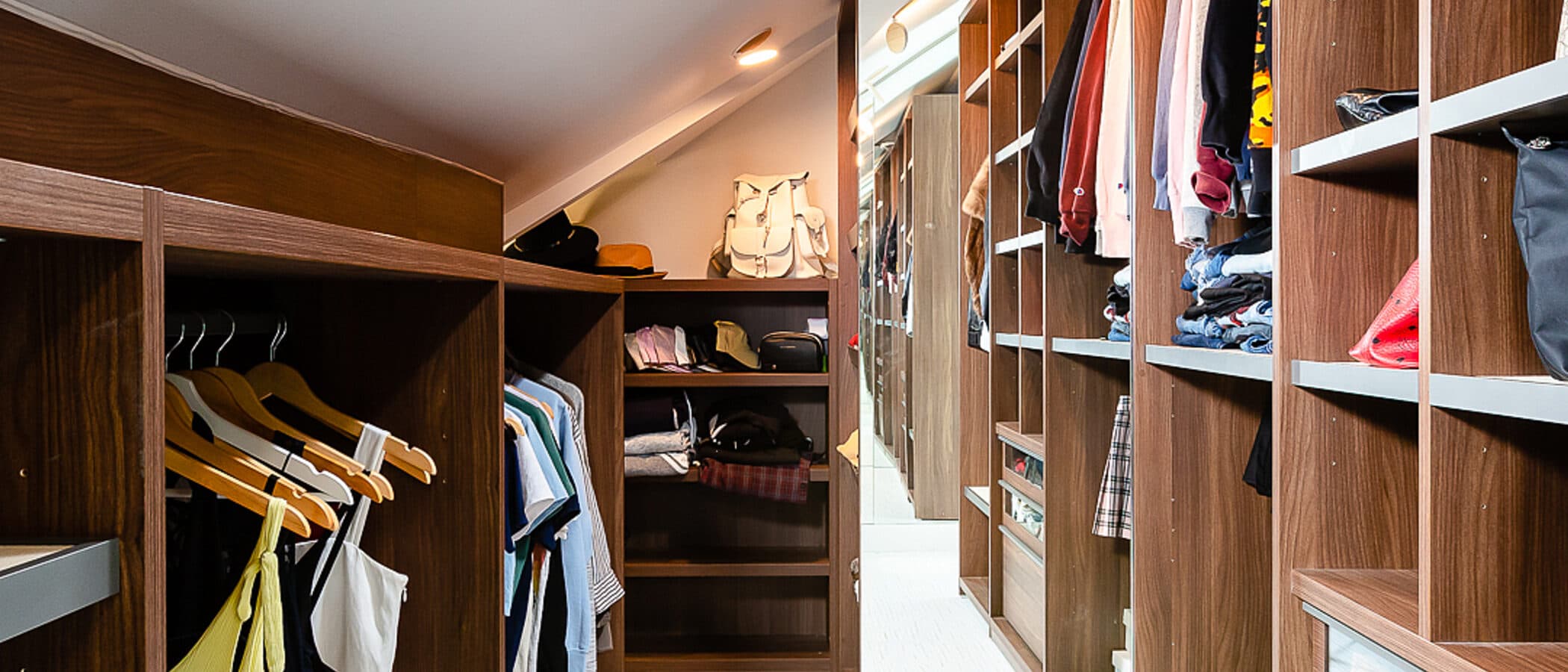 Walnut walk in closet with open hanging bays, built in drawers, and a mirrored end wall under a sloped ceiling