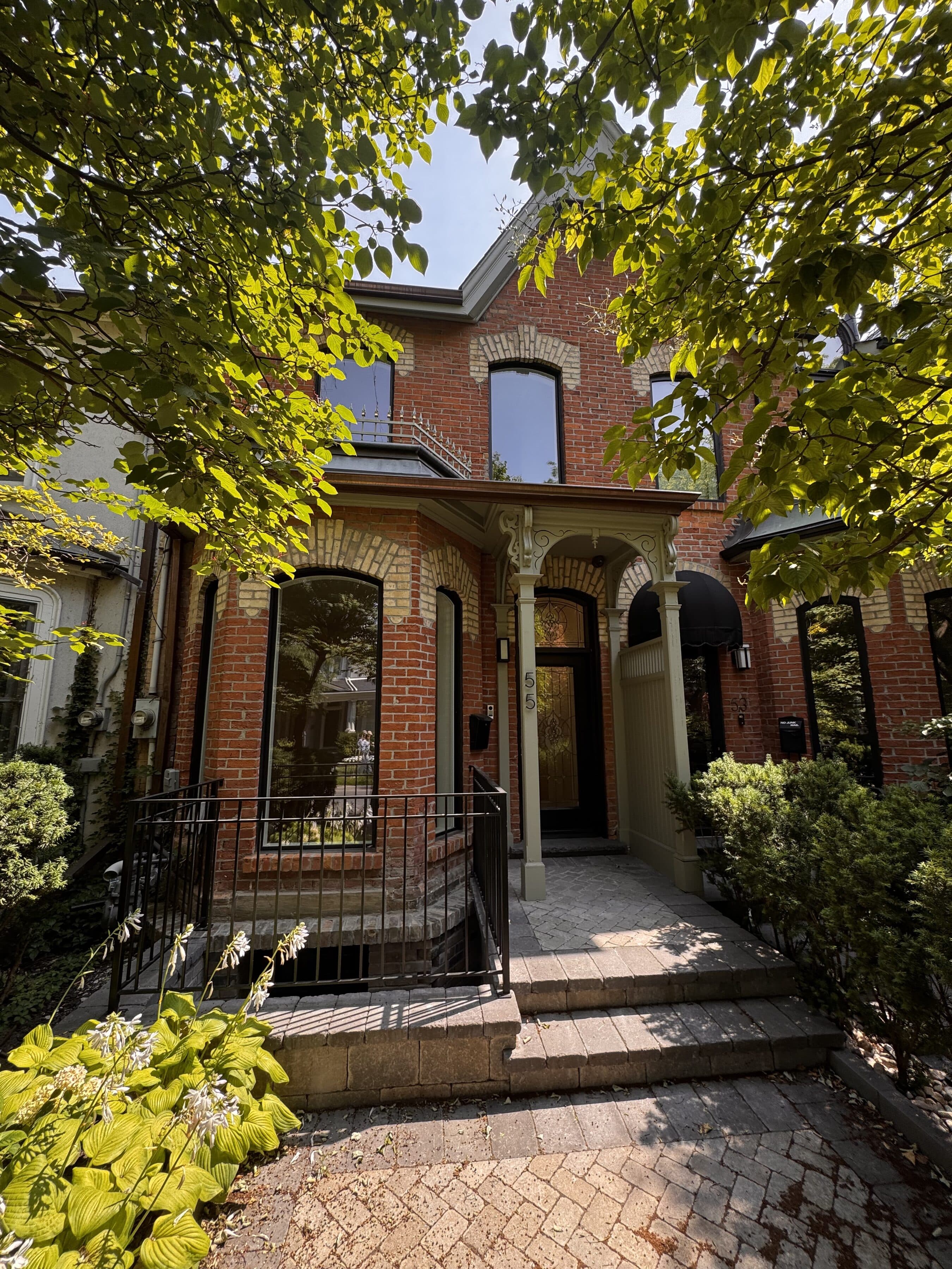 Red brick Victorian facade framed by mature trees, front entry beyond an iron gate