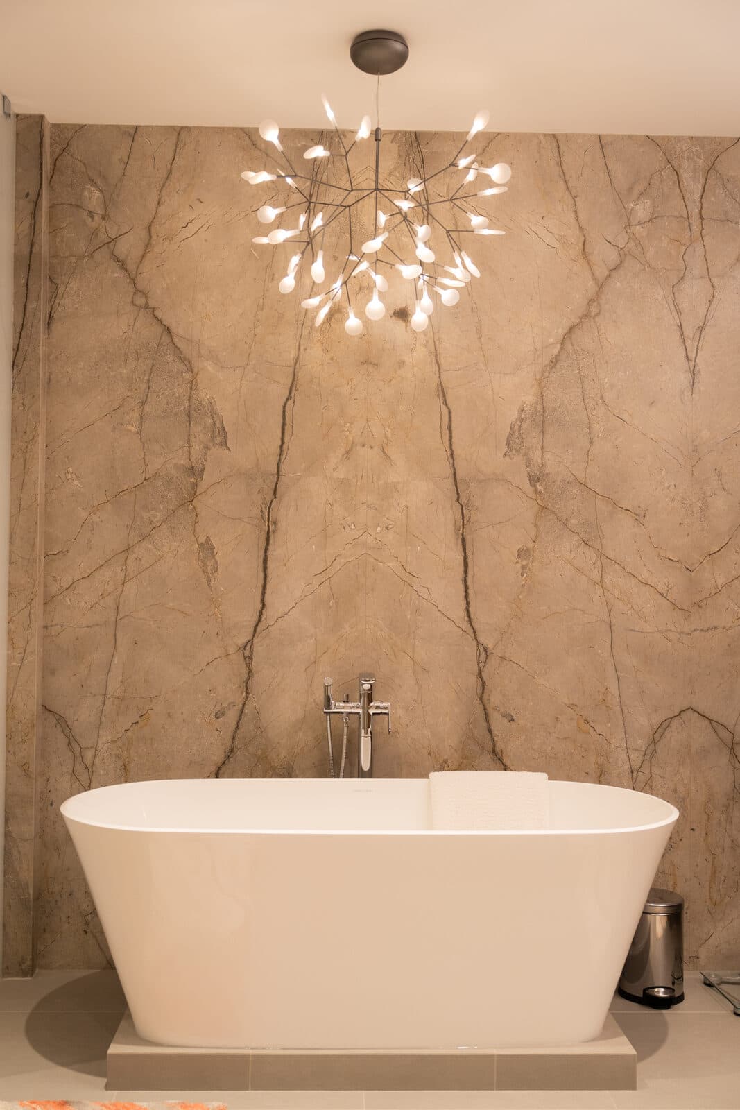 Freestanding tub beneath a branch form chandelier against book matched stone