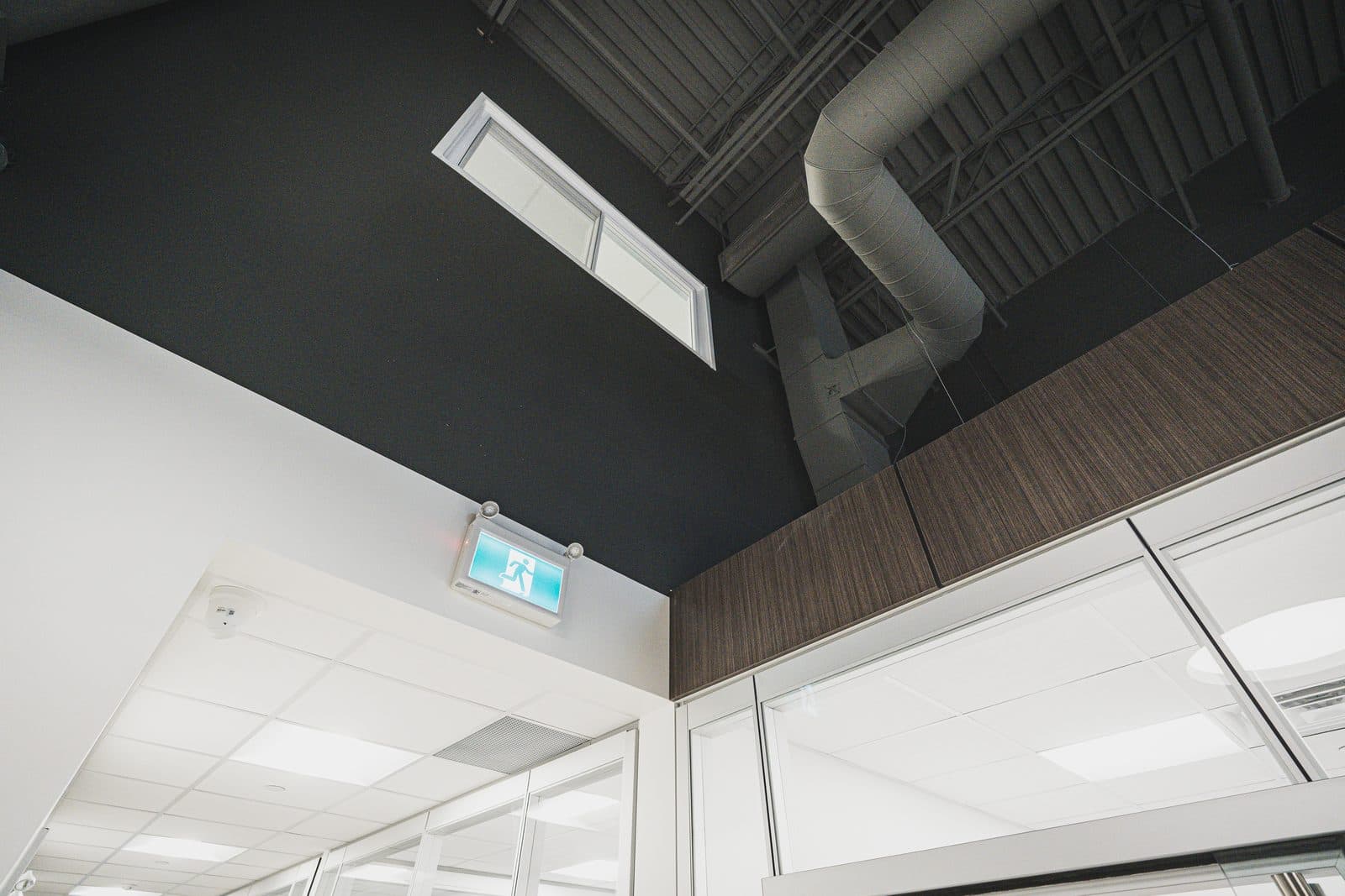 Black ceiling plane meeting white acoustic tile above glazed office fronts, with a clerestory window and spiral ductwork.