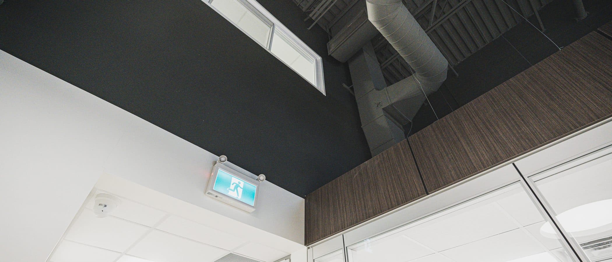 Black ceiling plane meeting white acoustic tile above glazed office fronts, with a clerestory window and spiral ductwork.