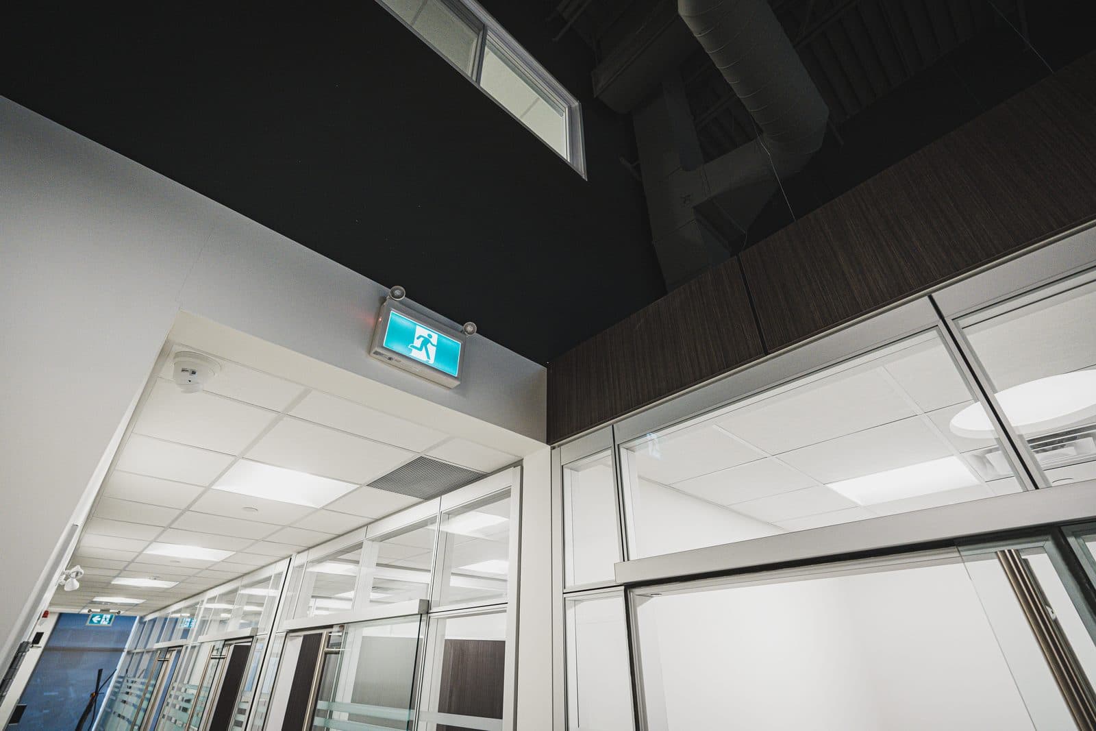Exposed ductwork and structure above a black ceiling plane, framing a glazed corridor of glass mounted offices below.