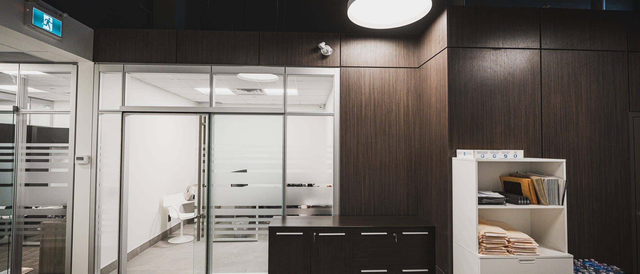 Small glass fronted meeting room with a dark wood credenza beneath pendant lighting and a striped privacy film door.