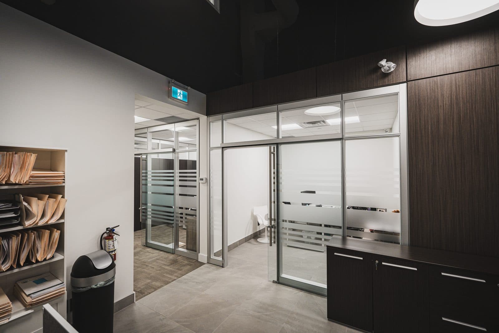 Corridor leading past a walnut millwork credenza toward a glass fronted meeting room and an illuminated exit sign.