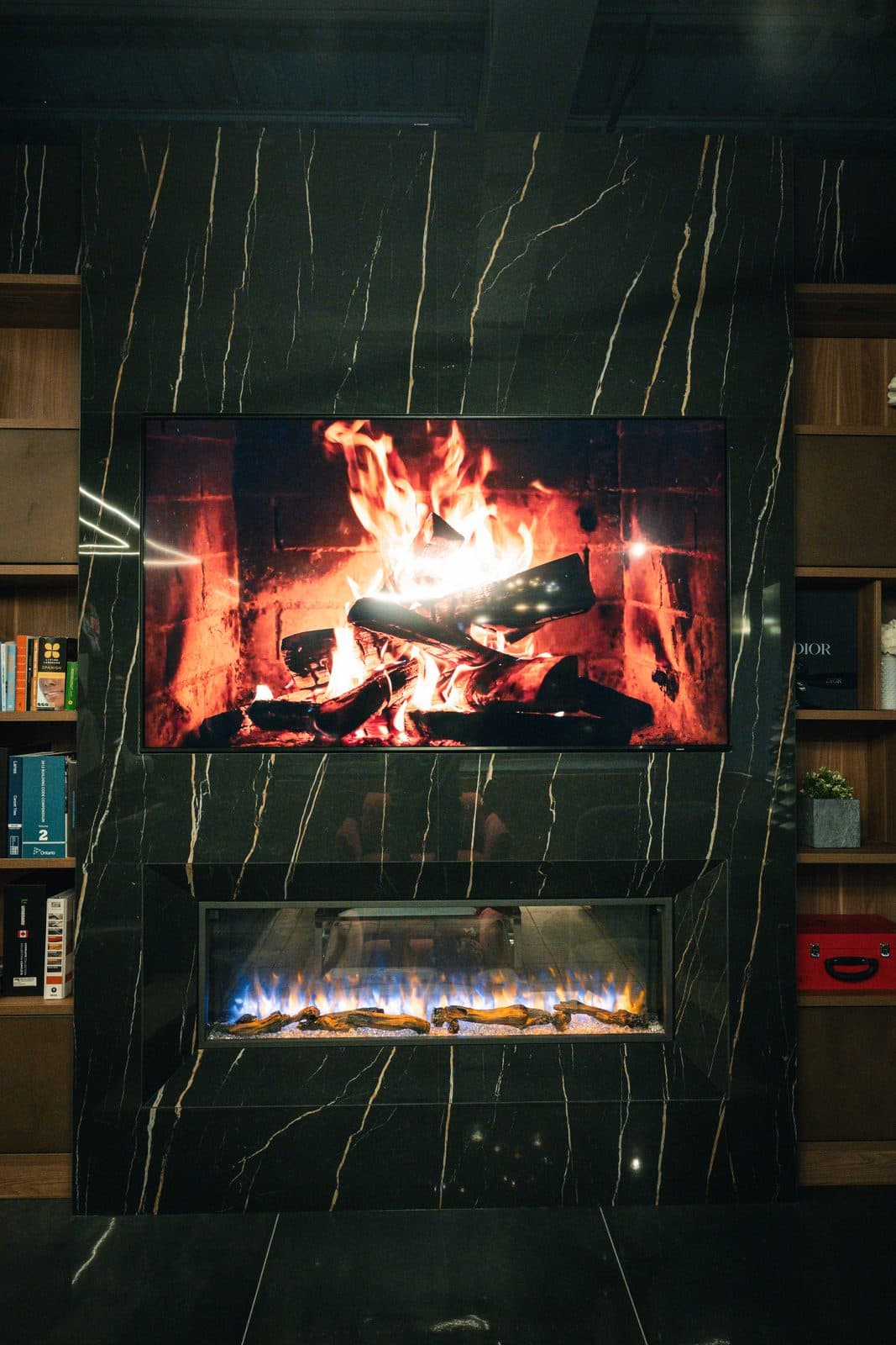 Linear gas fireplace framed in veined dark marble beneath a screen showing flame, flanked by walnut display shelving